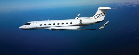 A Gulfstream G650 private jet in flight over an expanse of ocean, with a clear blue sky above.