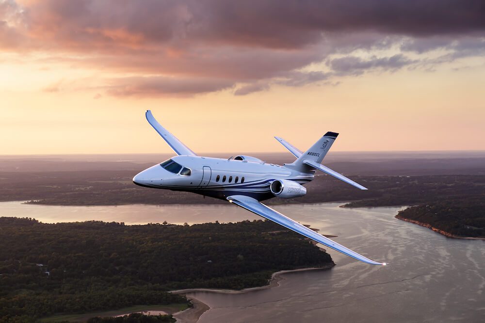 Cessna Citation Latitude flying over a large river and dark green trees with a pale-colored sky in the background.
