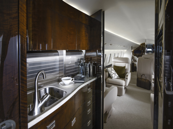 View from the Falcon 900LX galley into the forward cabin. The galley features dark polished wood cabinets, a white stone countertop and a stainless steel sink.
