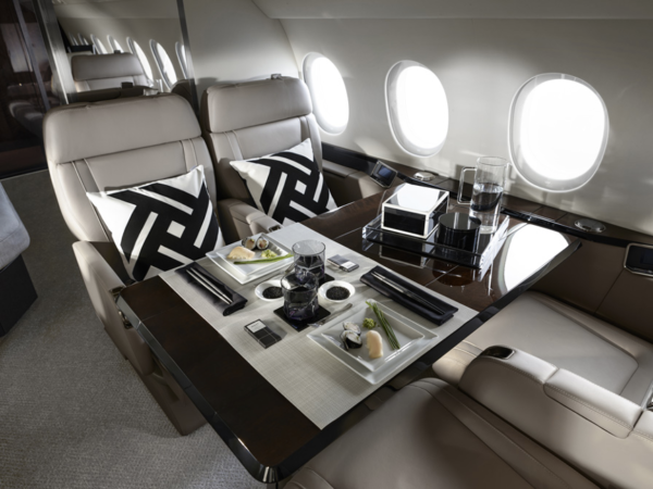 Falcon 900LX mid cabin with four beige leather upholstered seats arranged around a large, glossy black table. Two of the seats have pillows with a black and white geometric design on them, and an assortment of plates, glasses, cutlery and other objects.