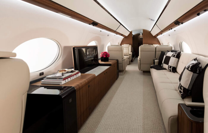 Private jet cabin with white walls, wood panel detailing and beige carpeting. A beige divan with pillows sits to the right side of the cabin, and a long wood credenza with magazines, flowers and an HD monitor sit to the left.