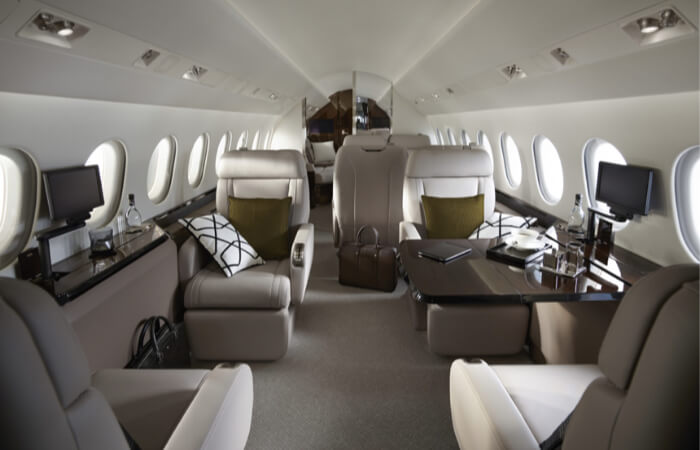 Private jet cabin with white walls and beige carpeting. Four executive-style seats with pillows and beige upholstery are arranged in a club configuration. There are HD monitors on both sides of the cabin, and the seats to the right face an extendable table. 