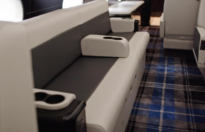 Closeup of bench-style seating with white and dark gray upholstery. The bench is divided by arm rests with cupholders, has storage compartments underneath and sits on a blue, gray and white plaid carpeting. 