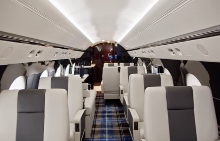 Private jet cabin with white walls and blue, gray and white plaid carpeting. Multiple rows of seating with white and dark gray upholstery are visible, and the wall at the back of the cabin is made of dark, glossy wood.
