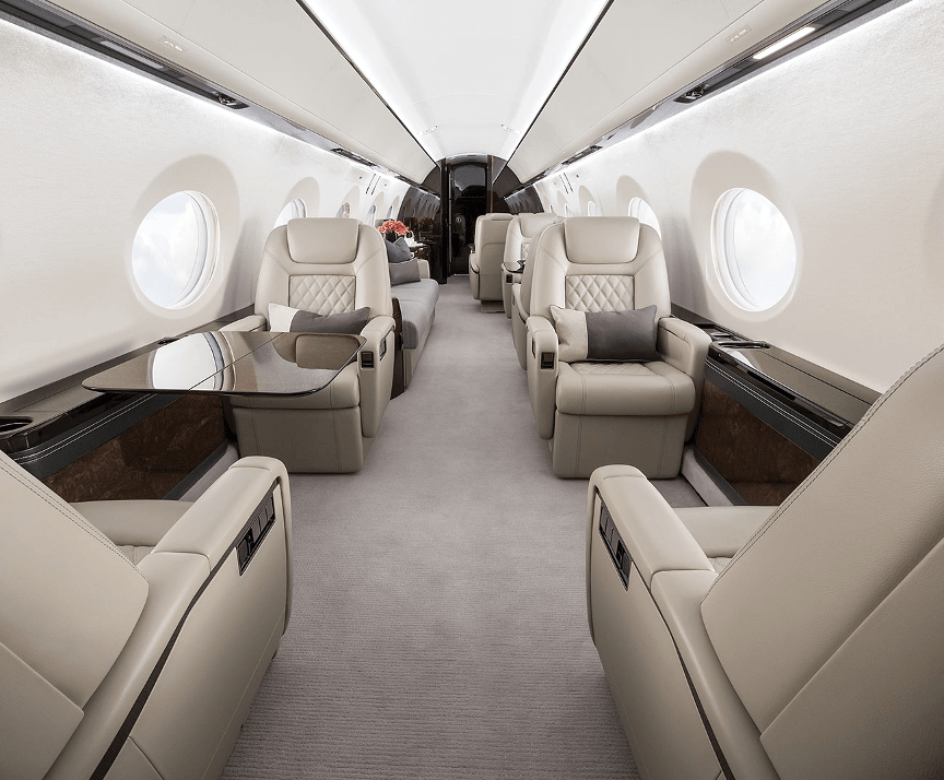 The Gulfstream G500’s forward cabin. The cabin features light gray carpeting, four seats upholstered in beige leather, and a dark wood table to the left.