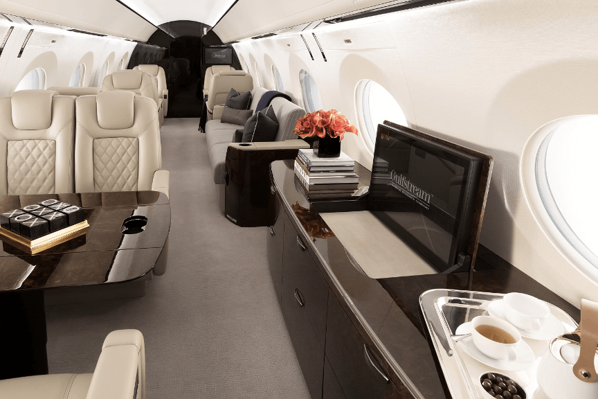 The Gulfstream G500’s mid cabin. To the right sits a dark wood credenza with an extended HD display monitor. To the left is a dark wood table and dual seats upholstered in beige leather on either side.