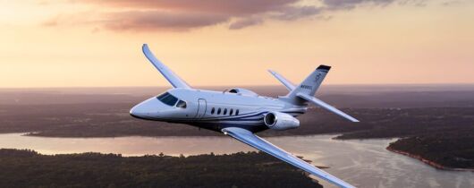 Cessna Citation Latitude flying over a large river and dark green trees with a pale-colored sky in the background.