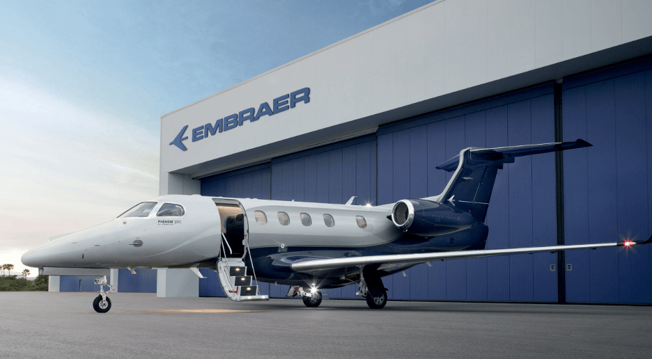 A white and dark blue jet sits with its airstairs lowered next to a gray and blue building marked with the name “Embraer.”