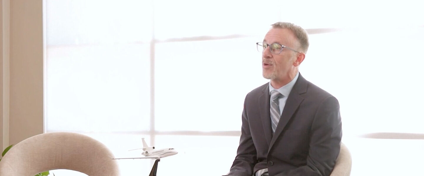 A man in a suit sitting in a brightly lit office, speaking, with a model of a private jet displayed on a table in the foreground.