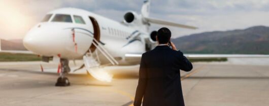 a man holding luggage and wearing dark clothing walks on a runways towards a private jet while holding a phone up to his ear